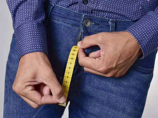 Closeup of a young man holding a piece of measuring tape that is popping up from the fly of his jeans, depicting the normal range of the penis